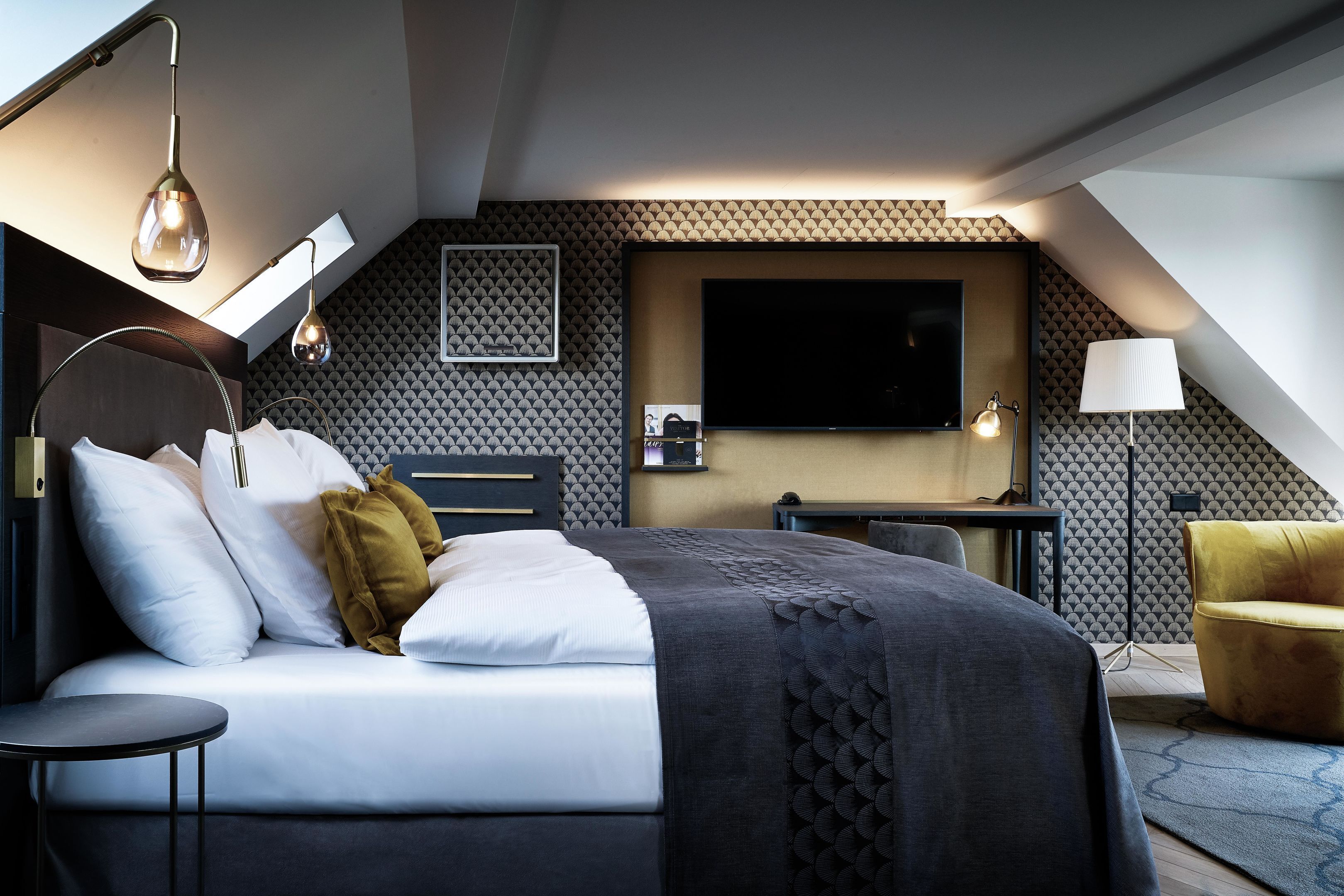 A modern hotel room with a bed, bedside table, lamp, and a TV mounted on the wall.