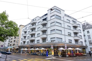 Ein mehrstöckiges Gebäude mit einer Bar im Erdgeschoss, Außenplätzen und einer Straßenbahn in der Nähe.