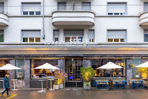 L’extérieur d’un hôtel, avec une enseigne indiquant son nom, des tables et des chaises à l’extérieur, protégées par des parasols, et des plantes en pot.