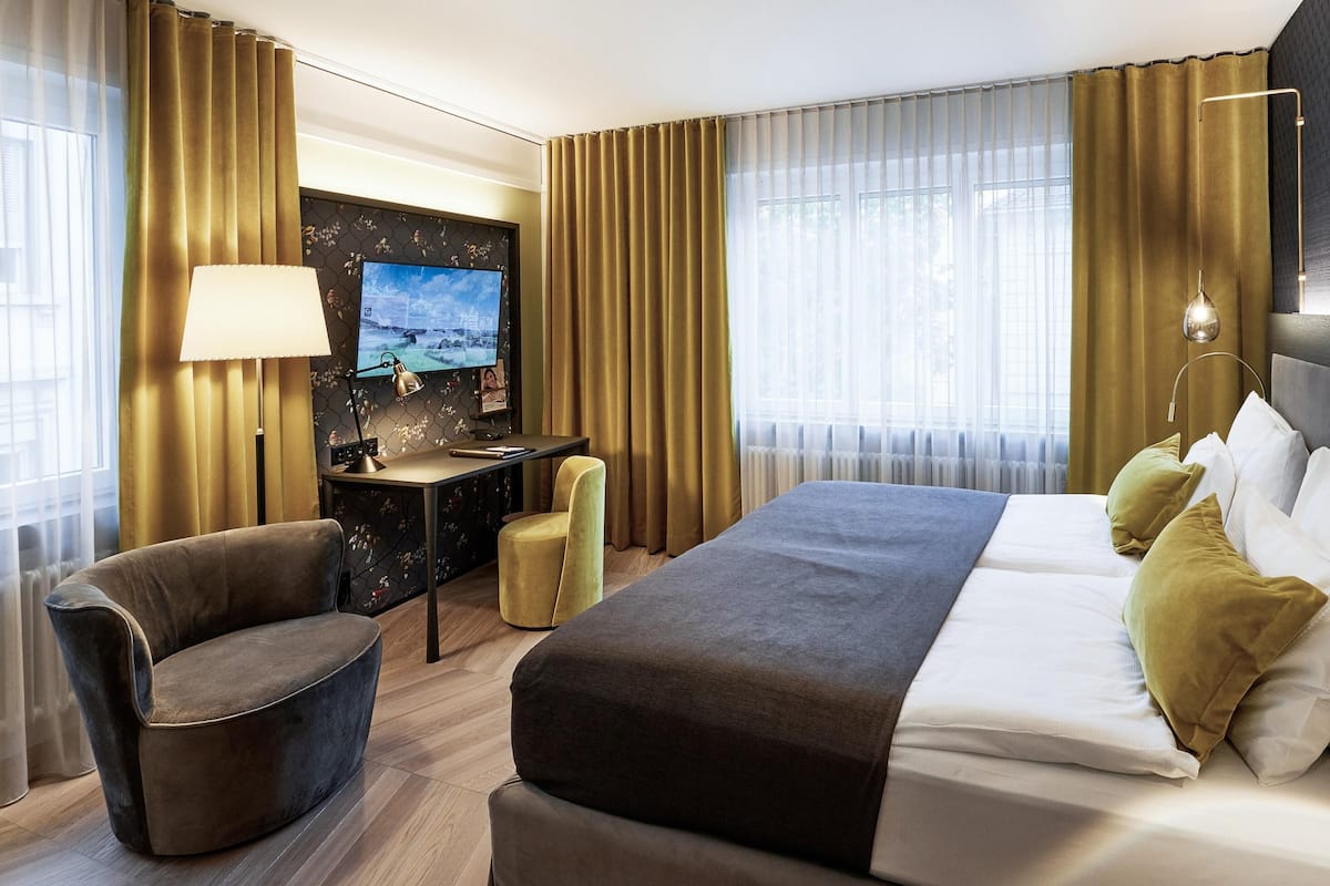 A hotel room with a bed, a desk with a lamp, a chair, and a TV on the wall.