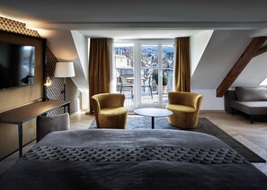 Habitación de hotel moderna con cama, dos sillones, una mesita, televisor y balcón con vistas.