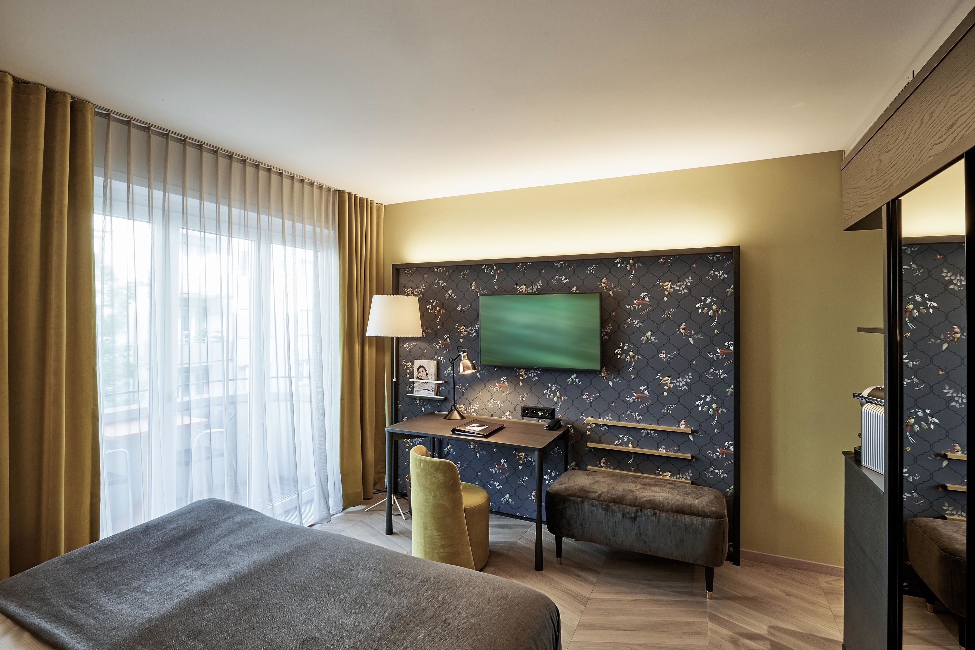 A hotel room with a bed, a desk, a chair, a TV mounted on the wall, and a window with curtains.