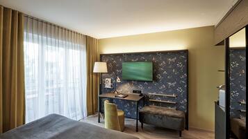 A hotel room with a bed, a desk, a chair, a TV mounted on the wall, and a window with curtains.