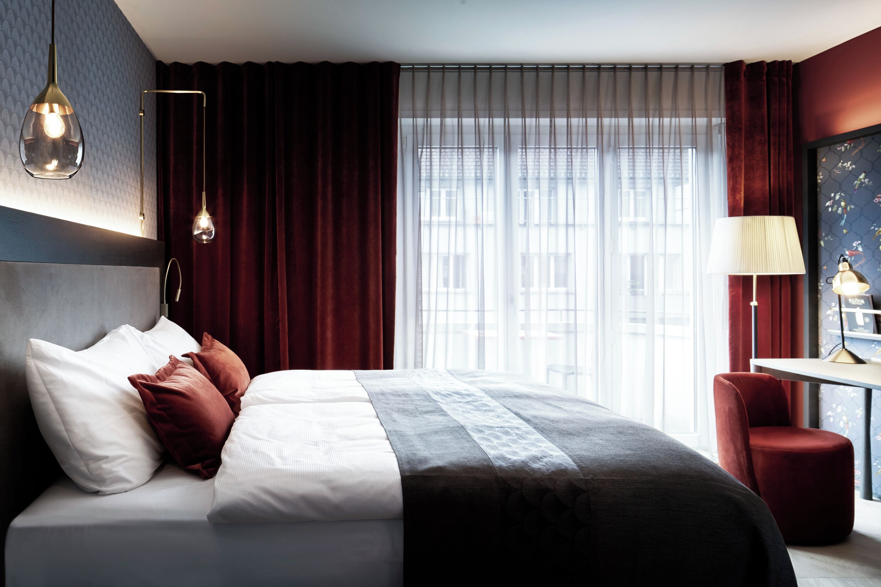 Ein Hotelzimmer mit einem großen Bett, einem Schreibtisch und Blick auf Gebäude durch transparente Vorhänge.
