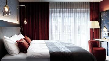 Ein Hotelzimmer mit einem großen Bett, einem Schreibtisch und Blick auf Gebäude durch transparente Vorhänge.