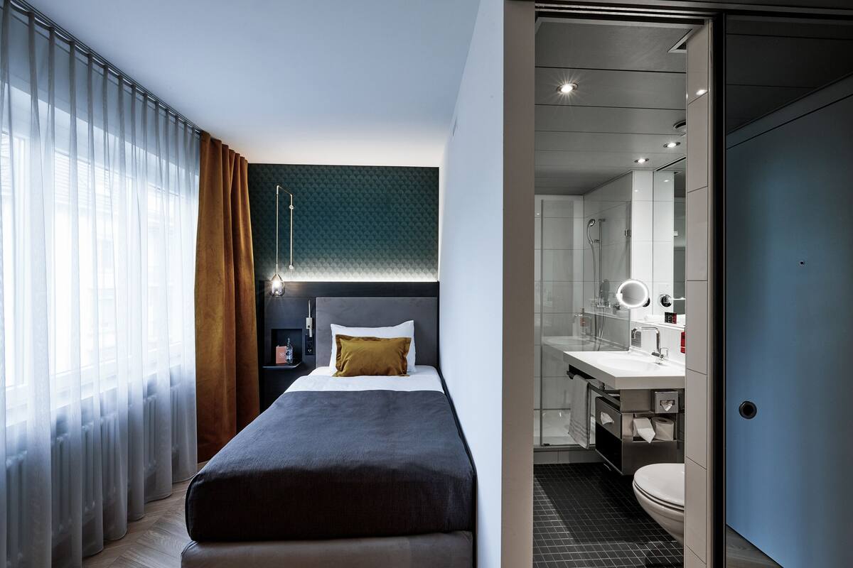 A modern hotel room with a bed, a bathroom with a mirror, and a view of the city.
