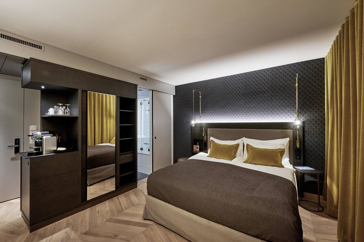 A modern hotel room with a large bed, a walk-in closet, a bedside table, and a headboard with integrated lighting.
