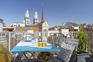A rooftop terrace with a table set for dining, a bench, and a view of historic buildings including a church with twin spires.