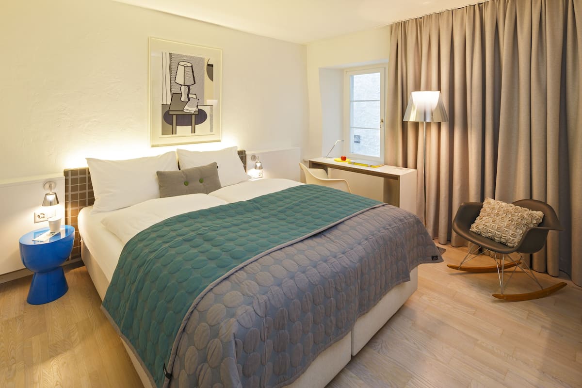 A modern hotel room with a large bed, a desk, a rocking chair, and a window with curtains.