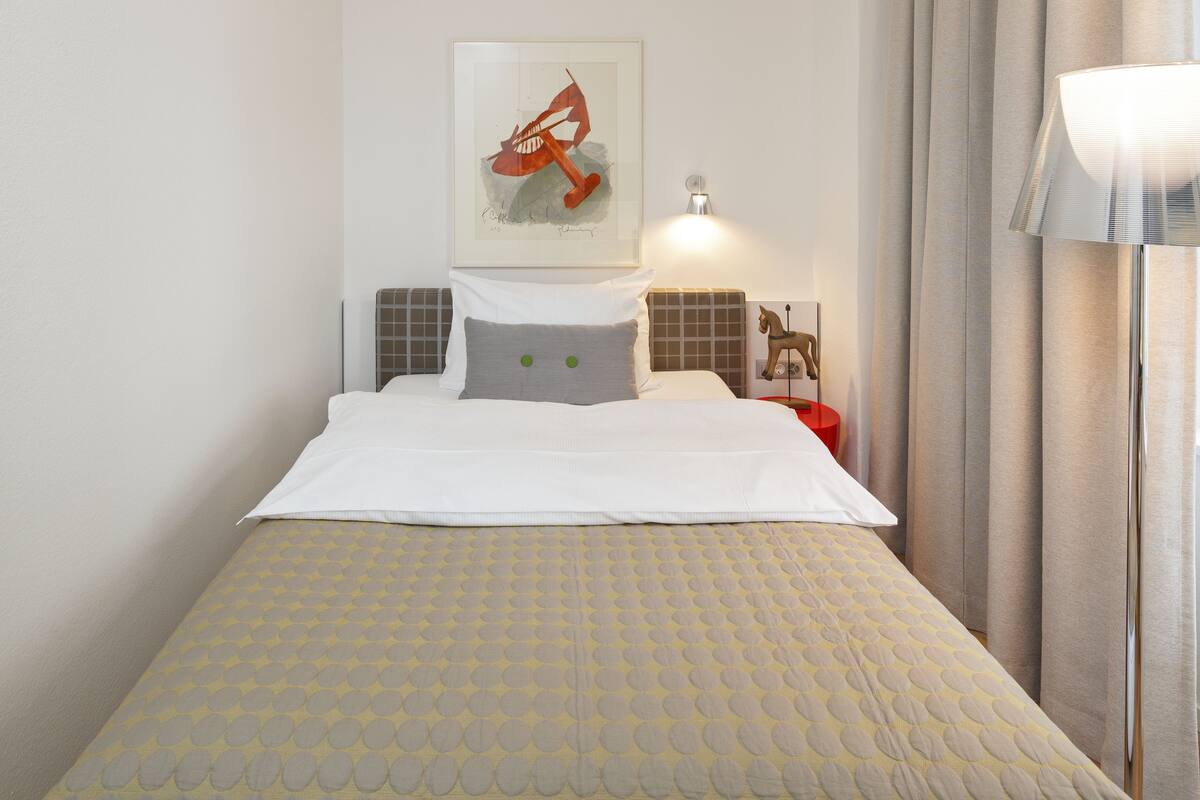 A neatly made bed with a patterned bedspread, a pillow with green buttons, and a framed artwork above it.