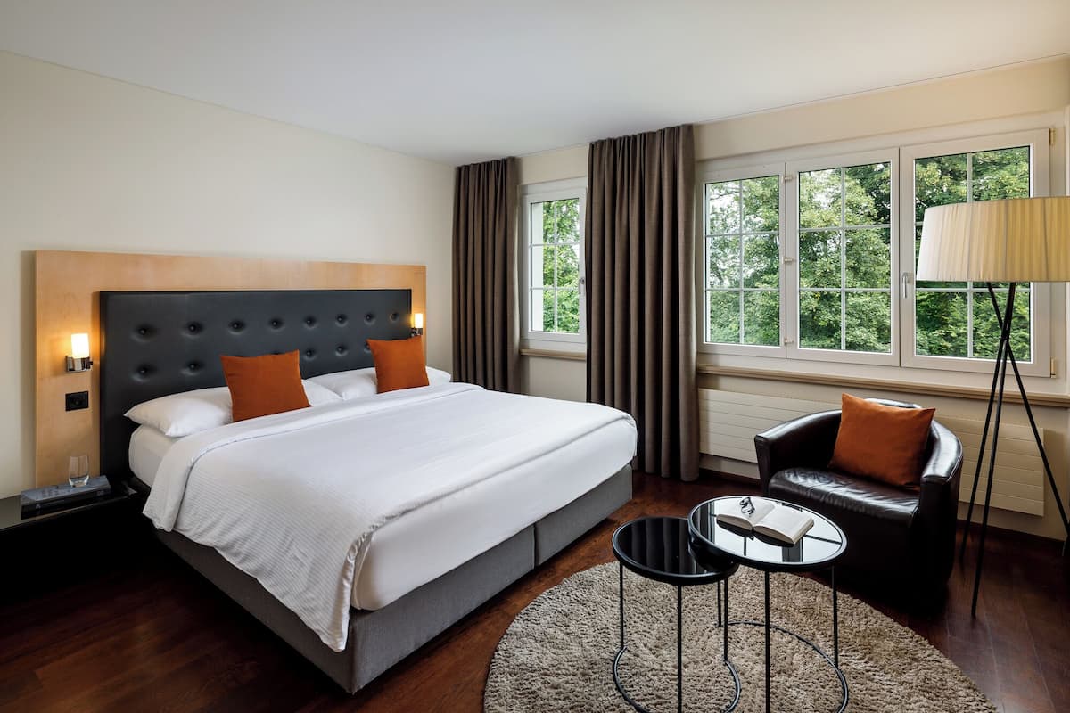 Une chambre d’hôtel avec un grand lit, une table de chevet, un fauteuil de lecture, une petite table et une fenêtre avec des rideaux.