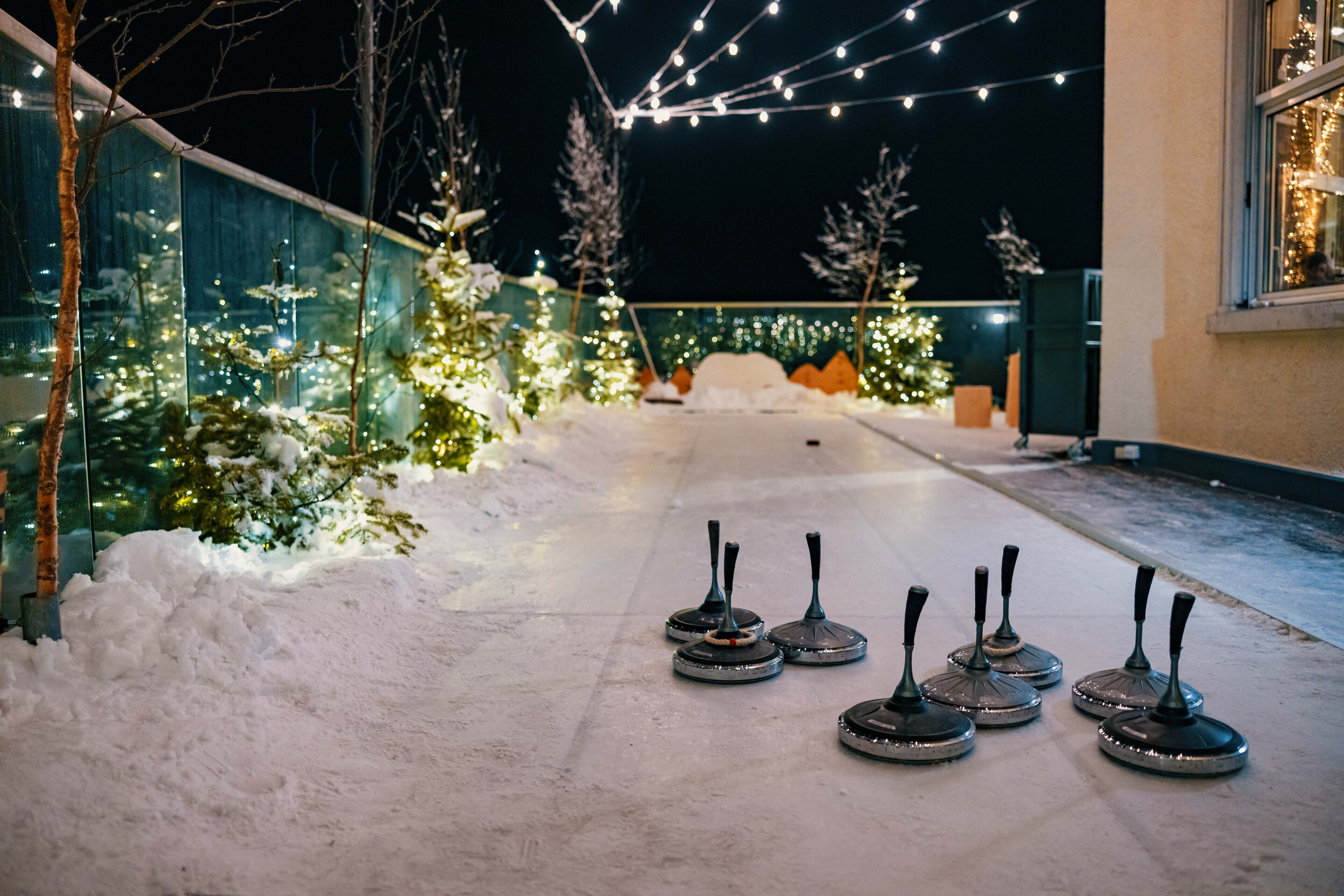 A snow-covered pathway with four curling stones arranged in a line, leading to a building with a glass facade and Christmas lights.