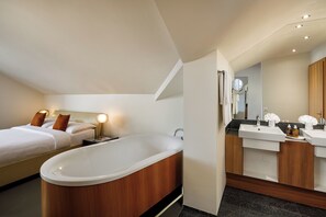 Una habitación de hotel moderna con una bañera grande, un dormitorio independiente con cama y lámparas de noche, y un baño con tocador y espejo.