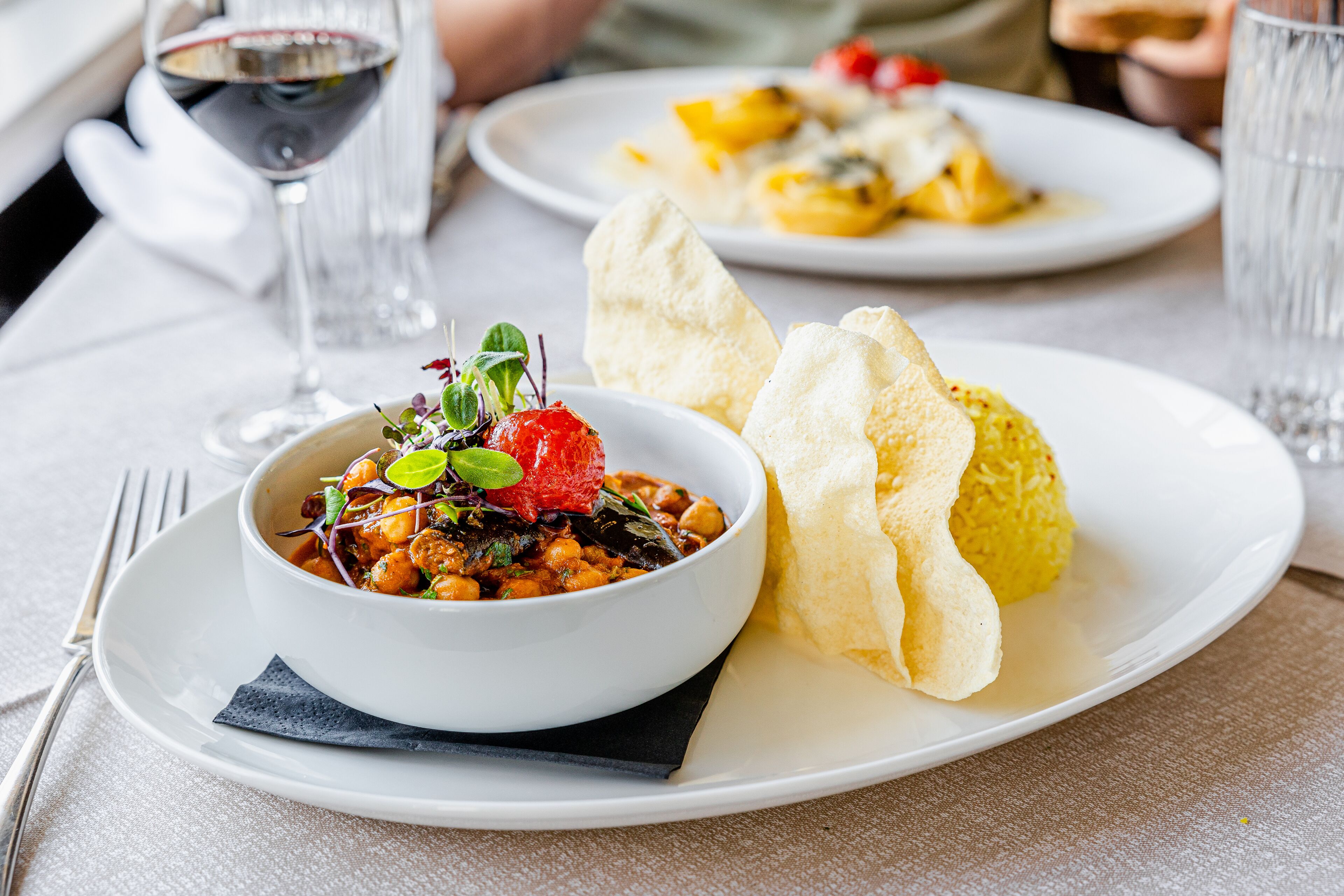 A dining table with a white plate containing a bowl of food, a glass of red wine, and a napkin.