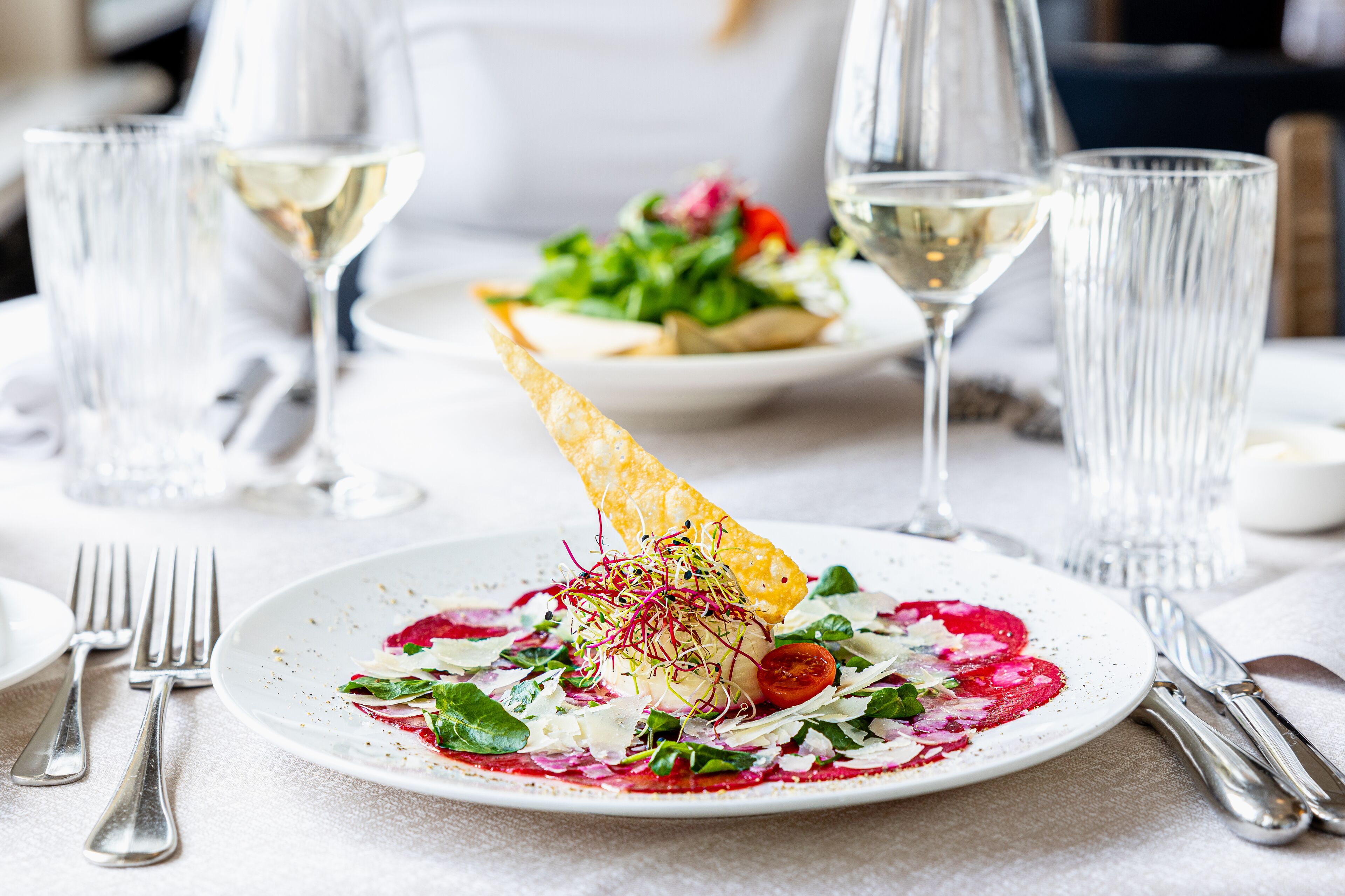 A dining table with a plate of food, a glass of white wine, and cutlery.