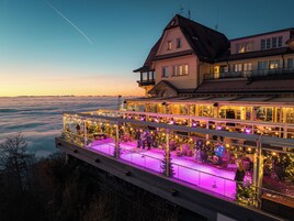 Un hotel con balcón que ofrece vistas a un paisaje montañoso, con zona de restaurante y asientos al aire libre con guirnaldas de luces.