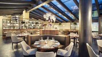 Um restaurante moderno com teto alto e claraboia, com mesas redondas preparadas para refeições, assentos confortáveis e uma exibição de utensílios de cozinha em prateleiras.