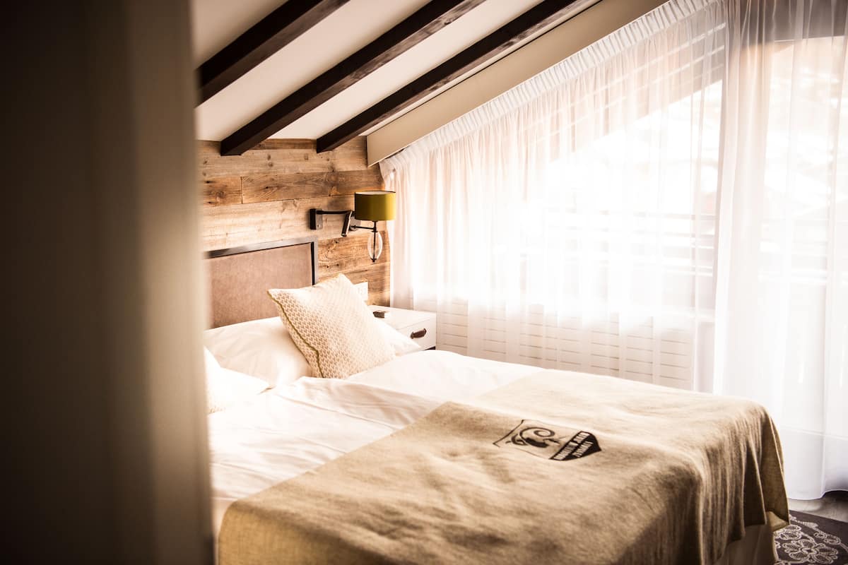 Una habitación de hotel con techo de madera, una cama con manta color beige, una mesita de noche con lámpara y cortinas traslúcidas.