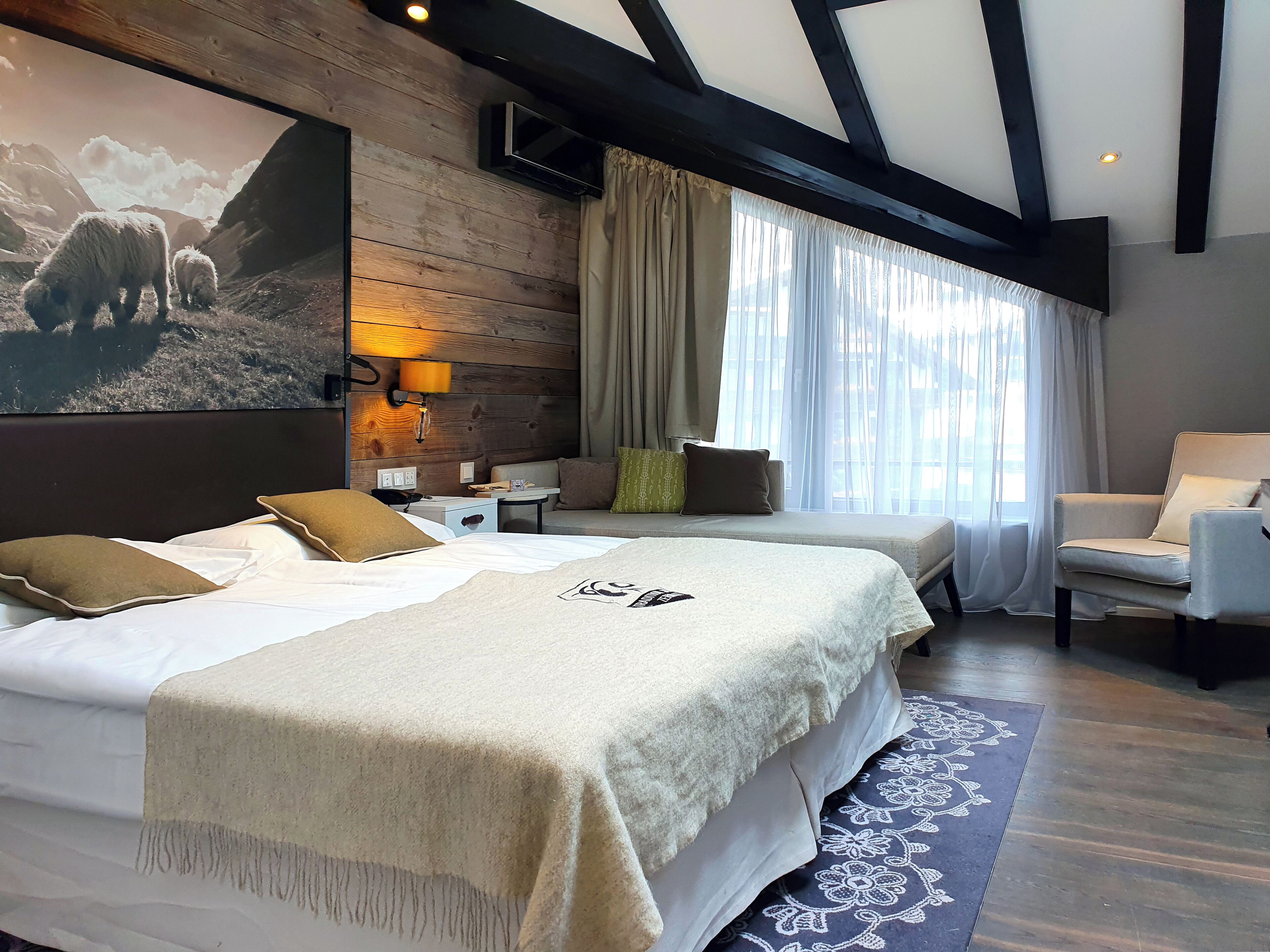 Quarto de hotel com duas camas, um sofá, uma cadeira, uma televisão e um quadro grande de ovelhas na parede.
