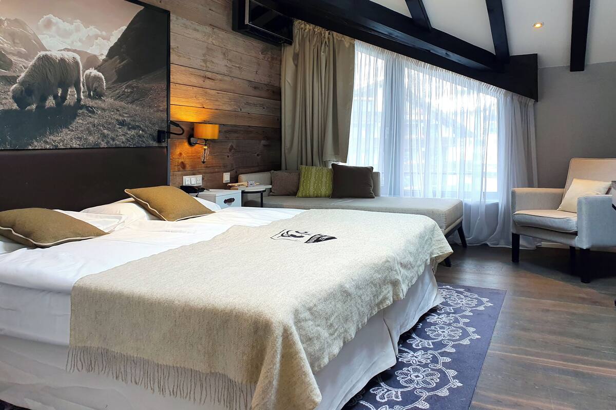 Habitación de hotel con dos camas, un sofá, una silla, un televisor y un cuadro grande de ovejas en la pared.