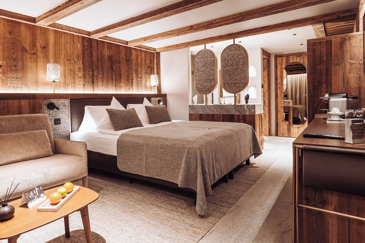 Una habitación de hotel moderna con techo de madera, una cama grande con edredón marrón, un sofá, una mesita con un jarrón y manzanas, y un escritorio con una cafetera.