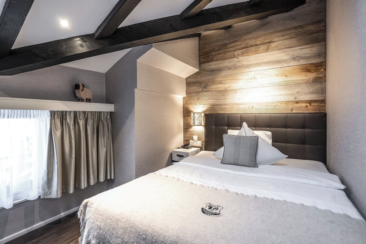 Un dormitorio con cabecera de madera, una mesita de noche con lámpara y una ventana con cortinas.