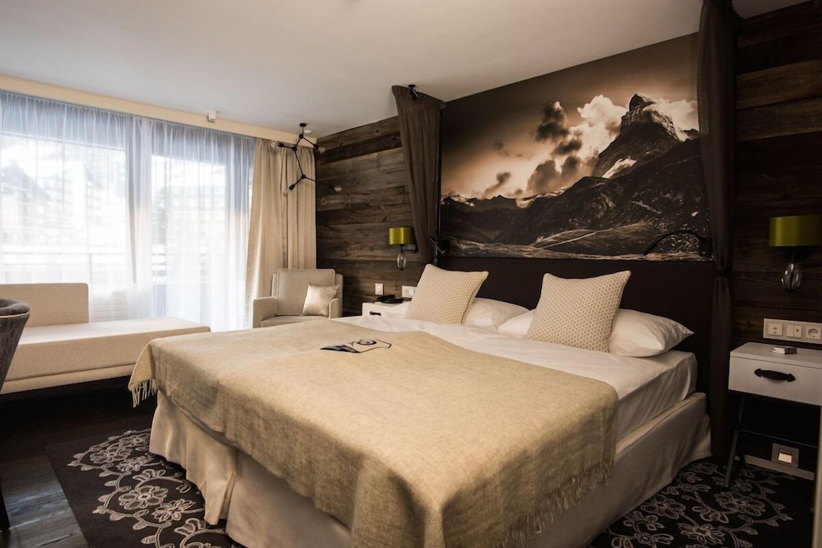 A hotel room with a bed, a chair, a nightstand, a lamp, and a large mountain painting on the wall.