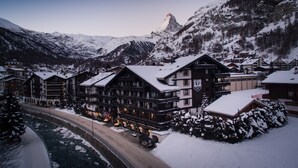 A snow-covered mountain town with a prominent hotel featuring multiple floors and balconies, surrounded by snow-covered trees and mountains. - Wellness Hotel Alpenhof (Zermatt)