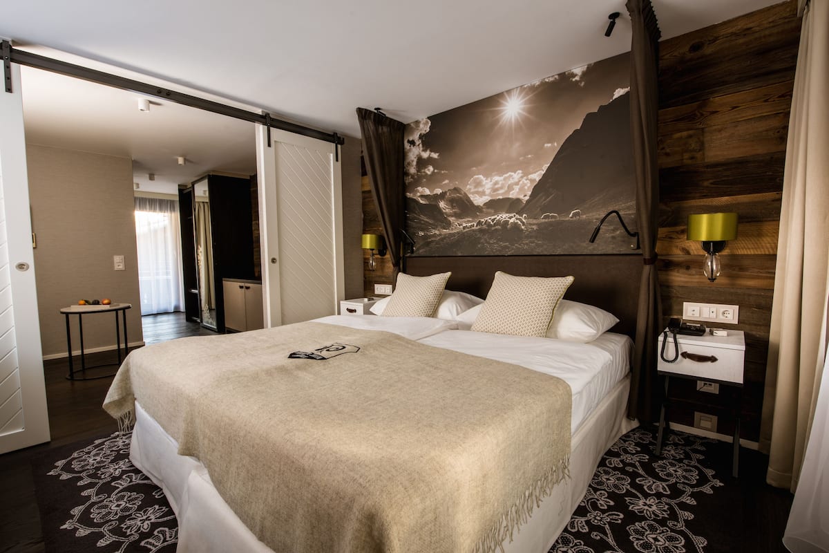 Habitación de hotel con una cama grande, una mesita de noche con lámpara, un teléfono en la pared y una puerta que da a otra habitación.