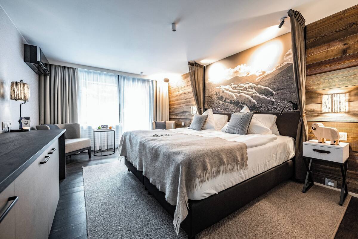 Habitación de hotel moderna con una cama grande, un escritorio y vistas a un paisaje montañoso por la ventana.
