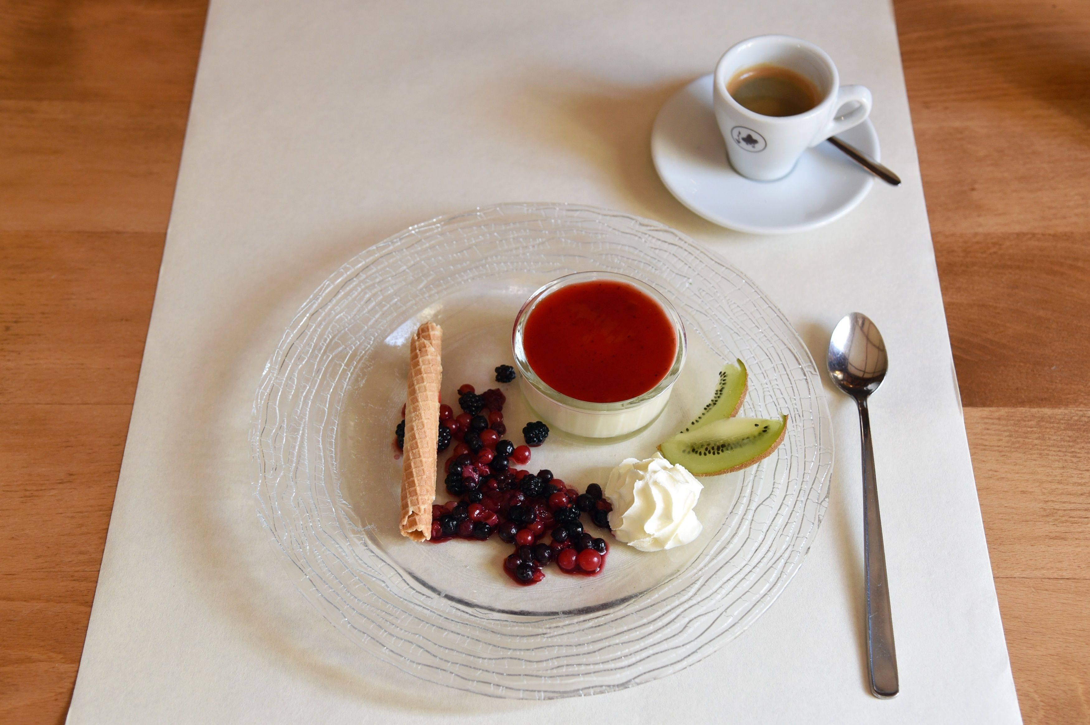 Ein Dessertteller mit Beeren, Schlagsahne und einem kleinen Glas rote Flüssigkeit, begleitet von einer Tasse Kaffee auf einem weißen Tisch.
