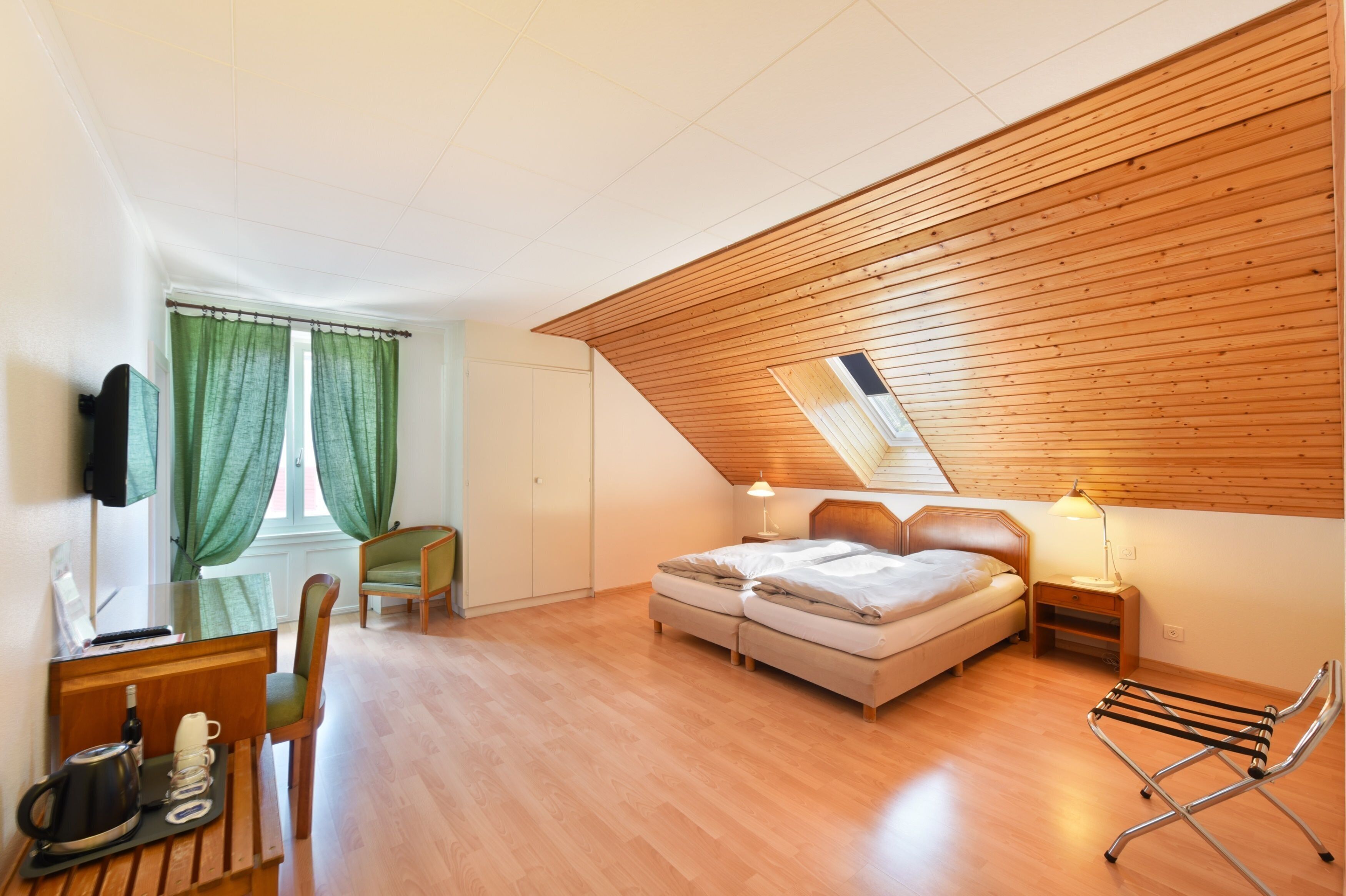 A bedroom with a lofted sleeping area, wooden ceiling, a desk with a chair, a TV, and a window with green curtains.