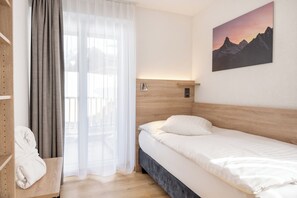 A hotel room with a bed, a wooden headboard, a bedside table with a phone, a window with sheer curtains, and a painting of mountains. - Hotel Bristol (Zermatt)