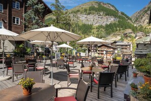 An outdoor patio with tables and chairs, surrounded by buildings and a mountainous backdrop. - Hotel Bristol (Zermatt)