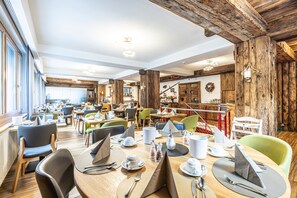A spacious dining area with multiple tables and chairs, wooden beams on the ceiling, and a view of the outdoors through large windows. - Hotel Bristol (Zermatt)