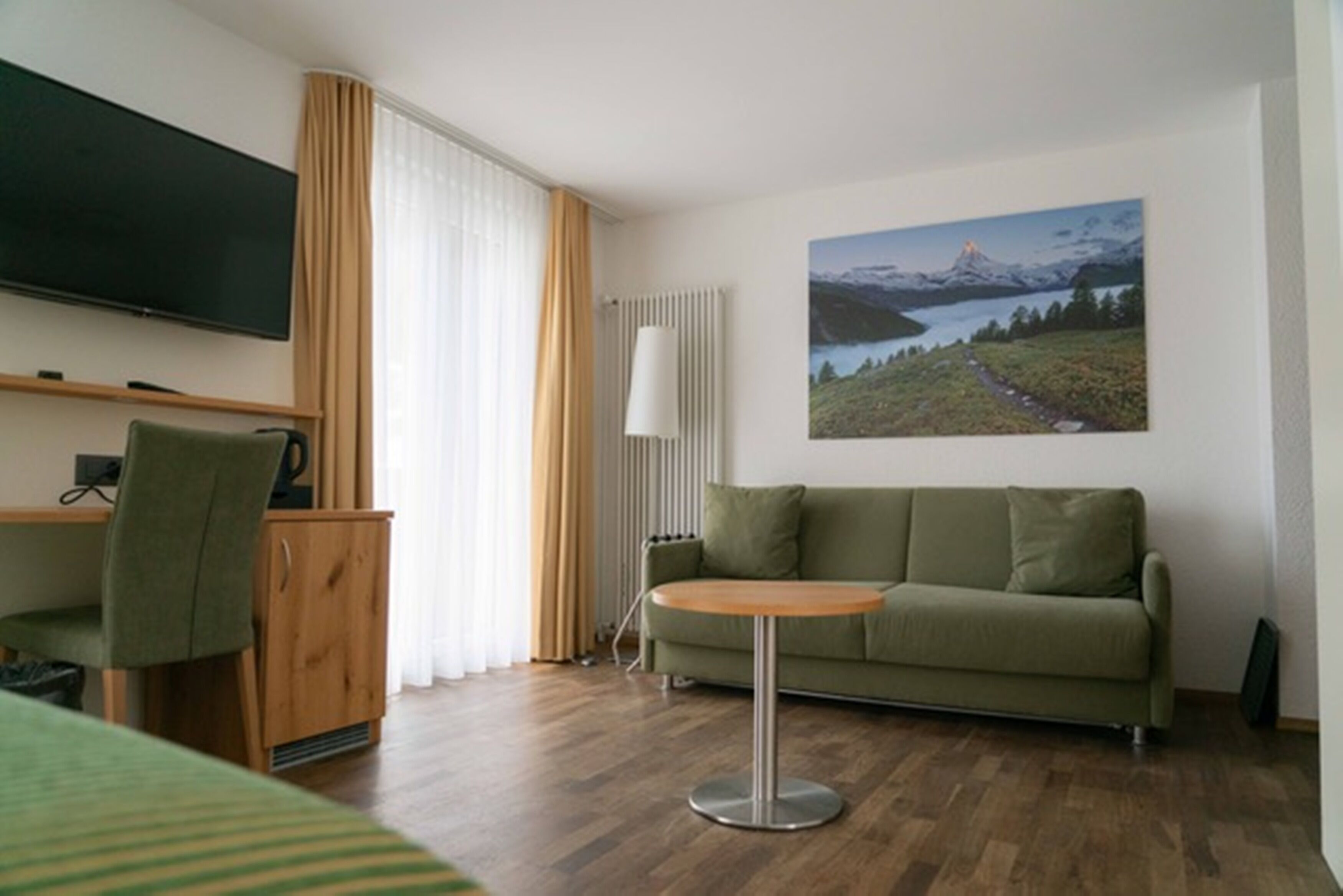 Een hotelkamer met een groene bank, een klein rond tafeltje, een aan de muur bevestigde televisie en een groot raam met gordijnen.
