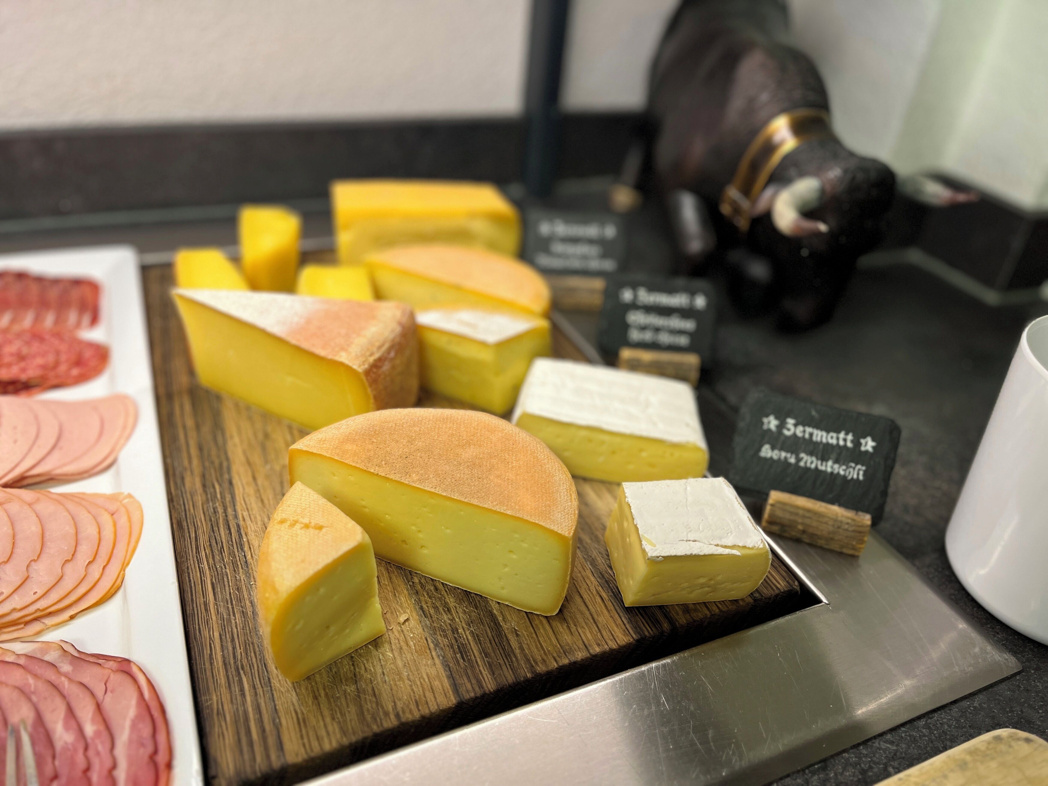 A selection of cheeses on a wooden board with labels, a knife, and a cup on a counter.