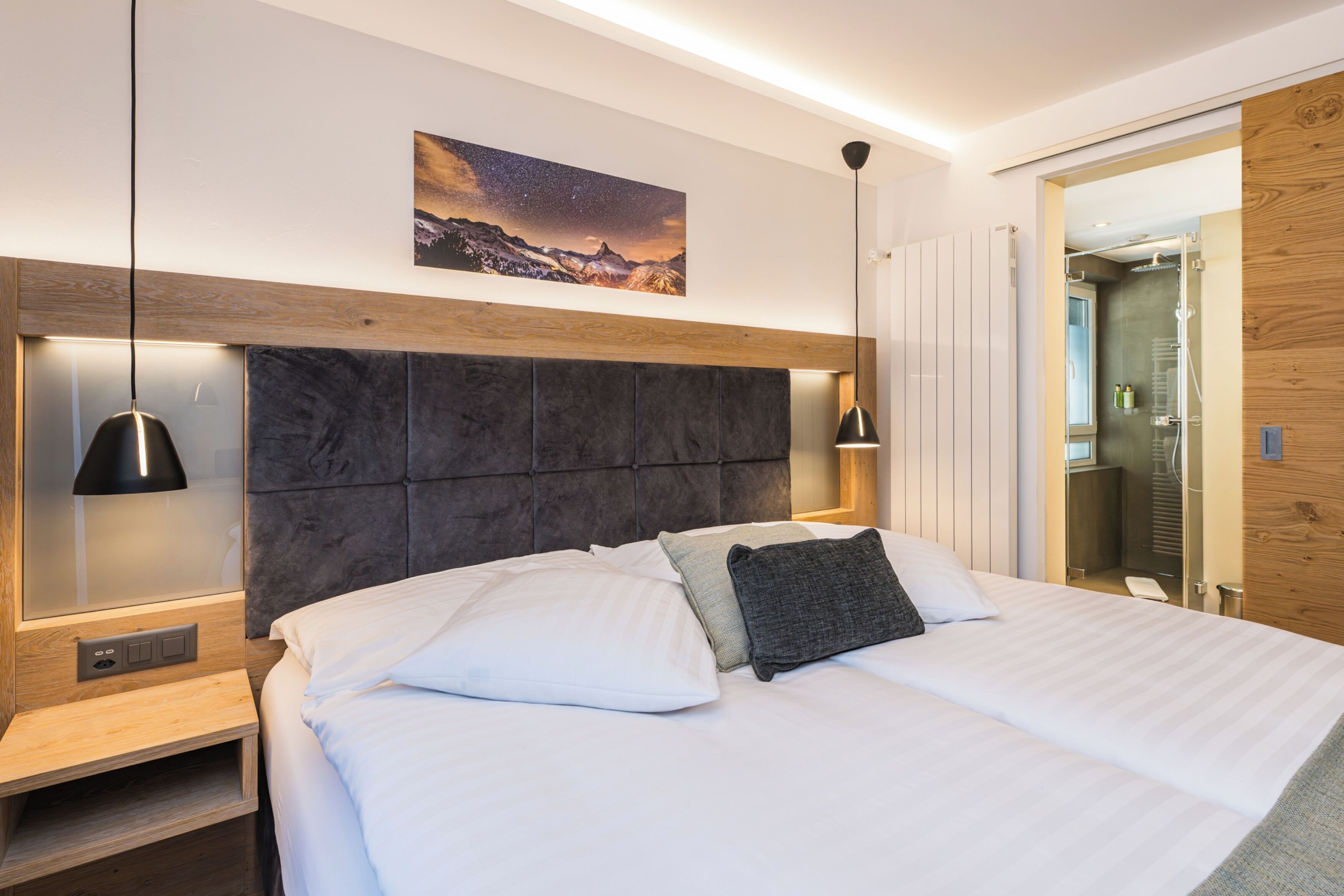 A modern hotel room with a large bed, a headboard with a wooden panel, a nightstand, a wall-mounted lamp, and a painting of a mountain landscape.