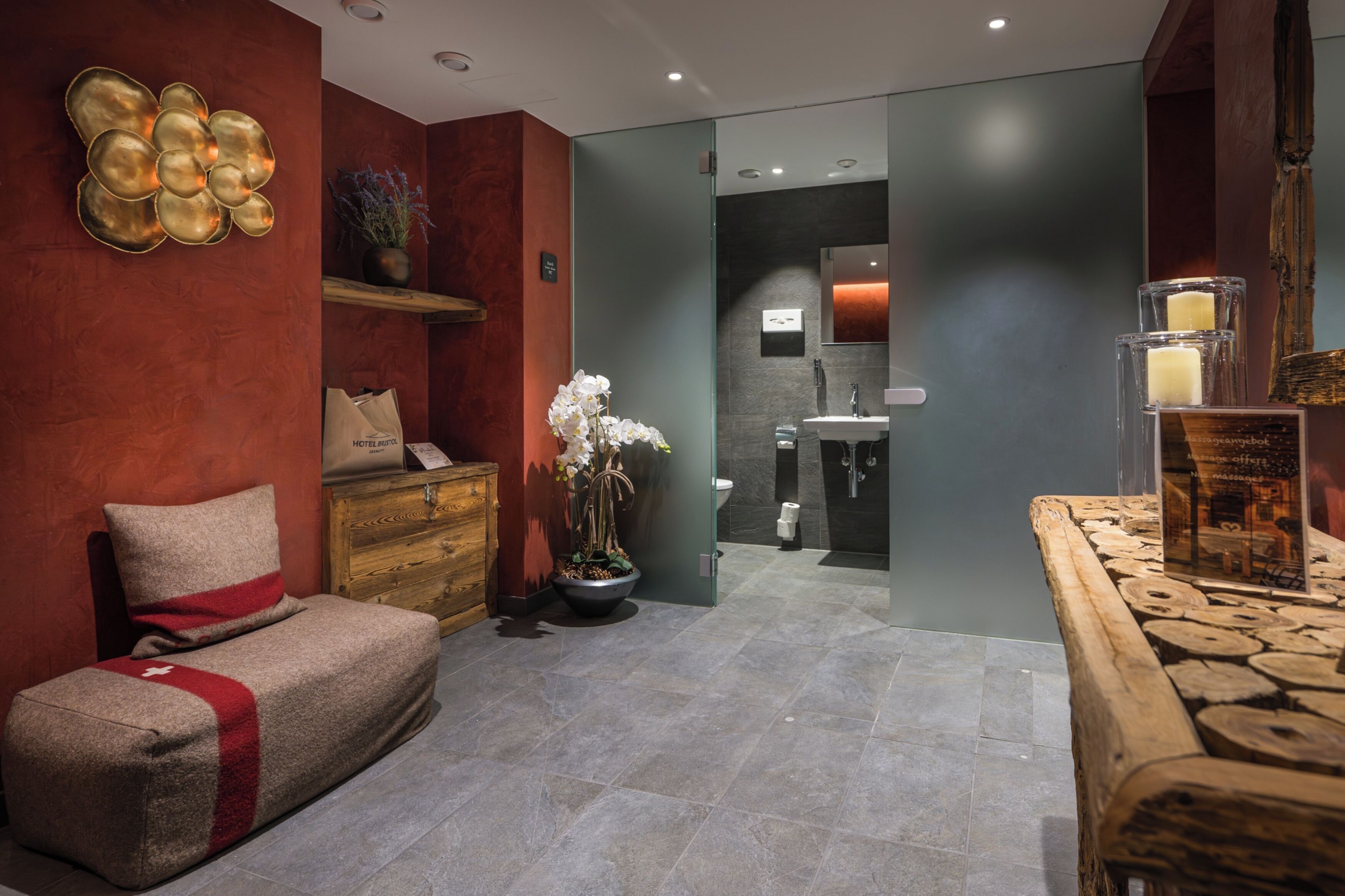 Habitación de hotel moderna con un sofá, un aparador de madera, una planta en maceta y un baño visible a través de una puerta de vidrio.