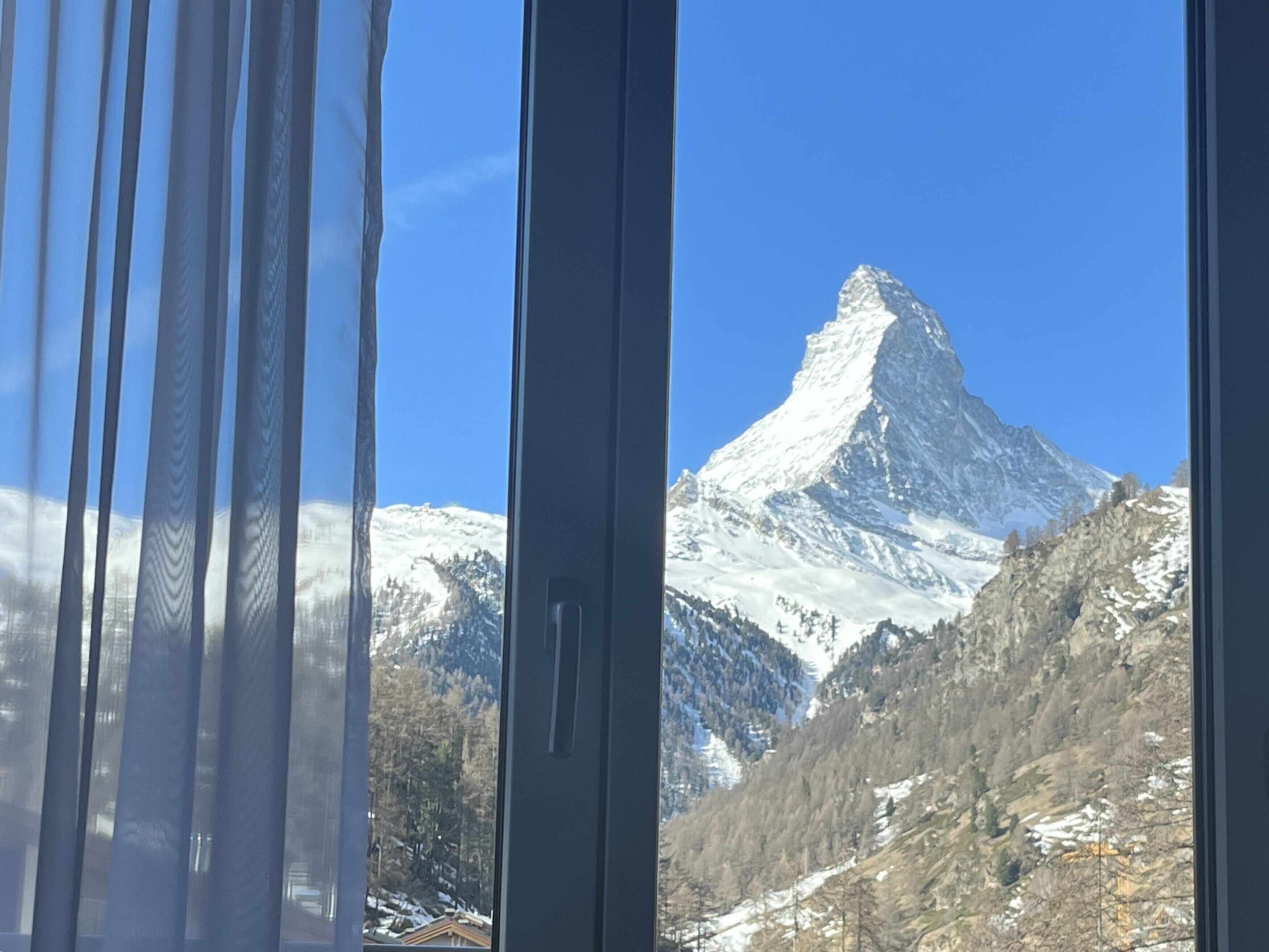 A mountain view through a window with curtains, featuring a snow-covered peak.