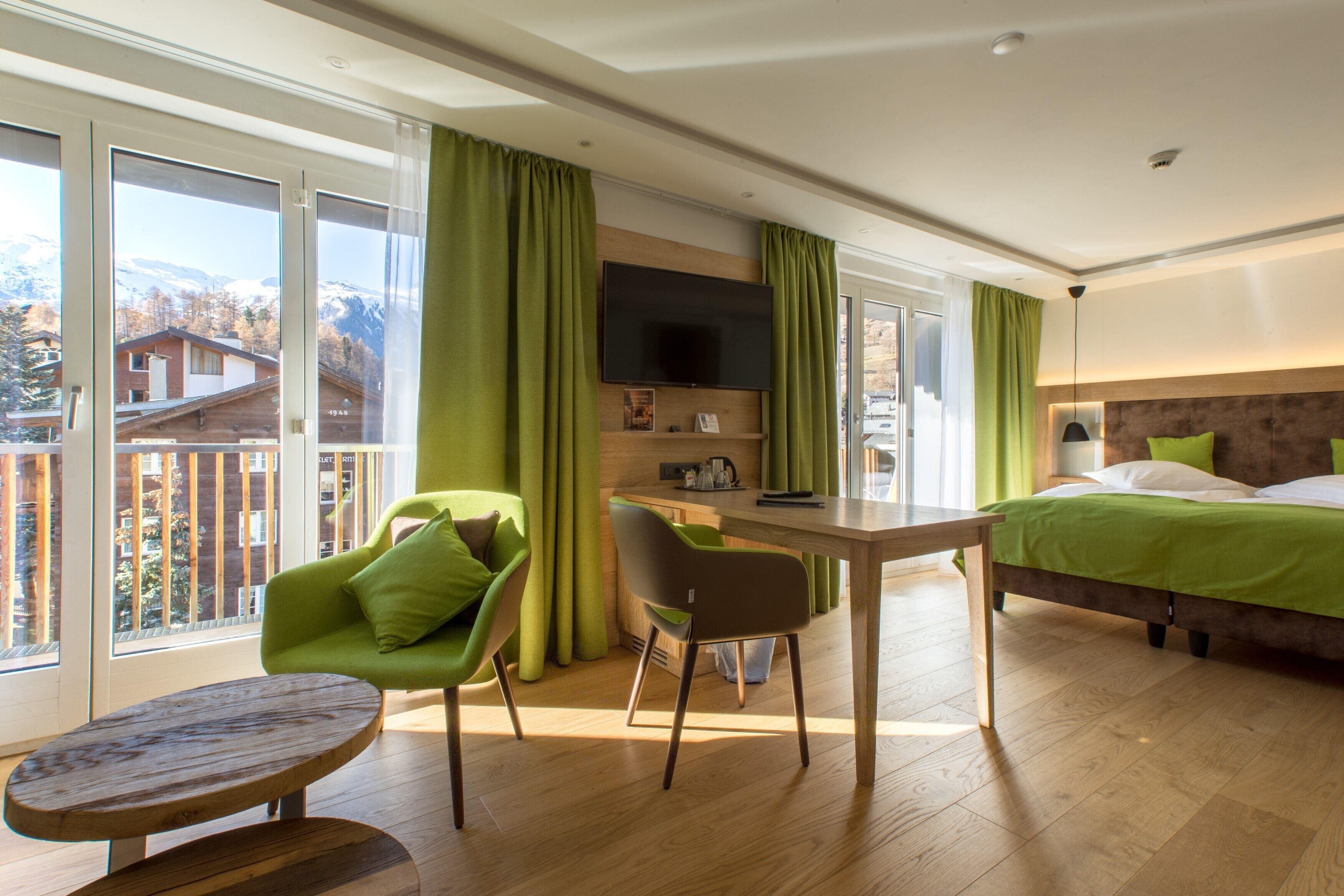 Ein modernes Hotelzimmer mit einem Holztisch, einem grünen Stuhl, einem Bett mit grüner Bettwäsche, einem Fernseher und großen Fenstern mit Vorhängen.