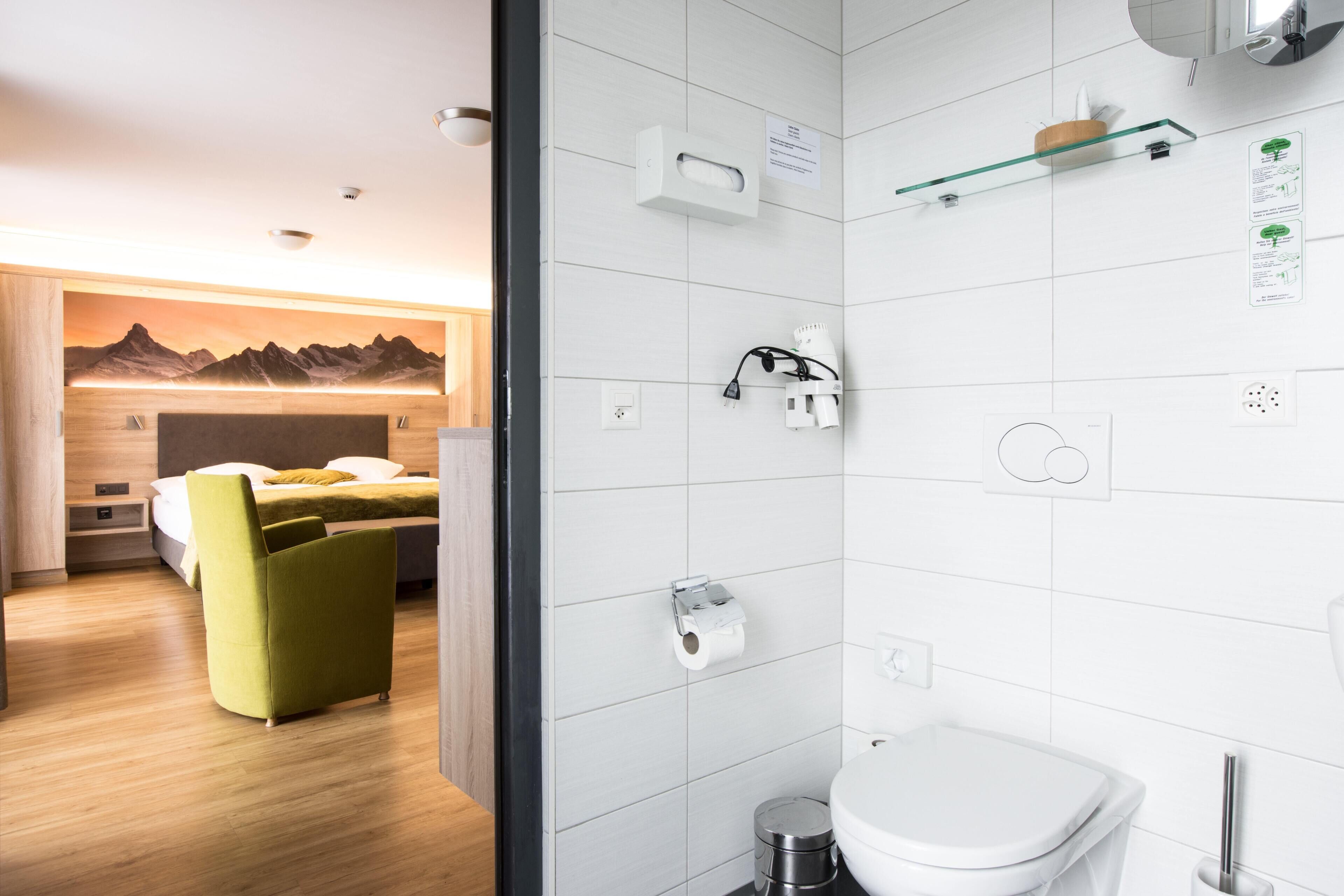 A hotel room with a bed, a green chair, a bathroom with a toilet, a mirror, and a shower.