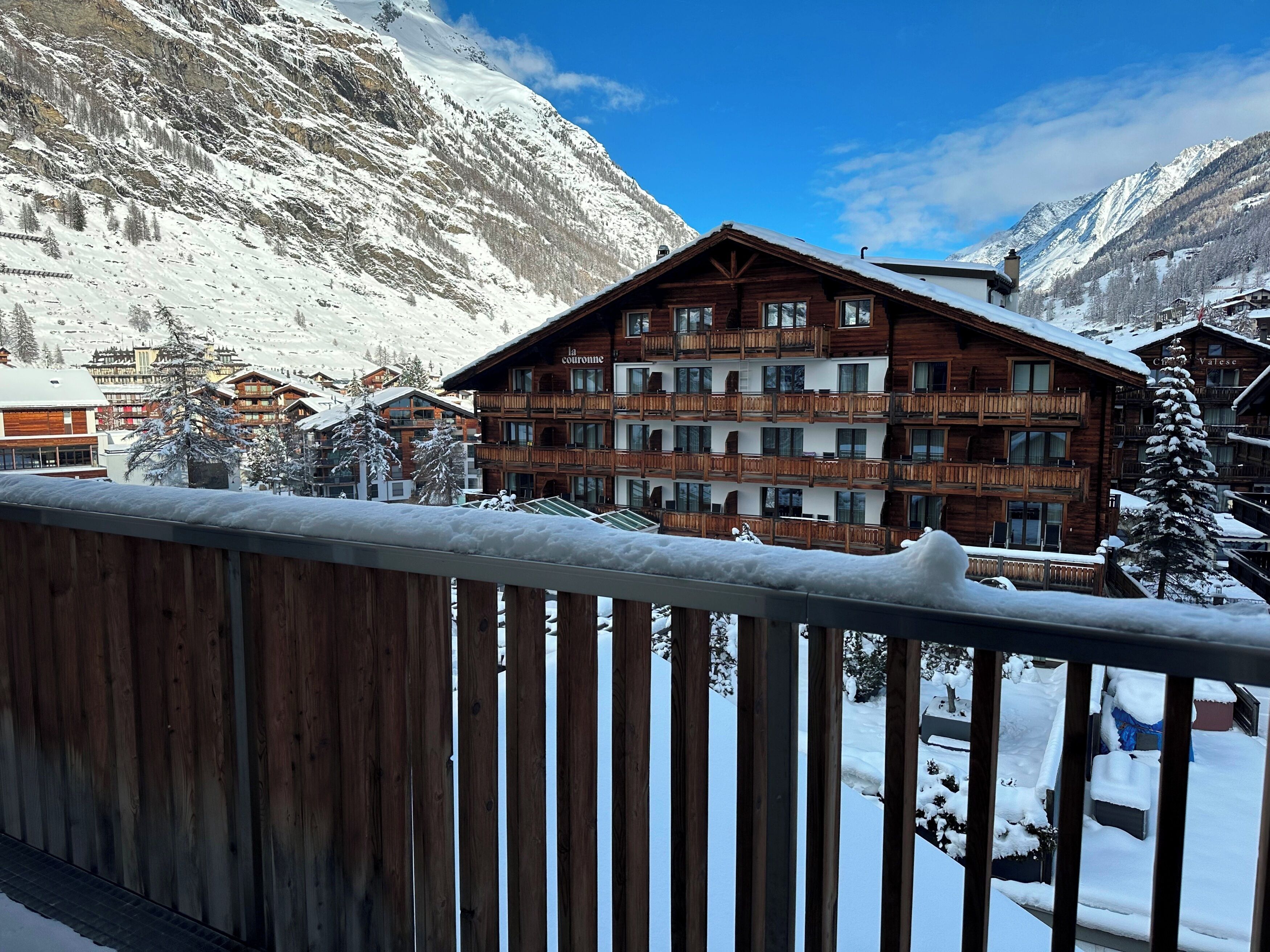 A wooden chalet with multiple balconies, surrounded by snow-covered mountains and other buildings.