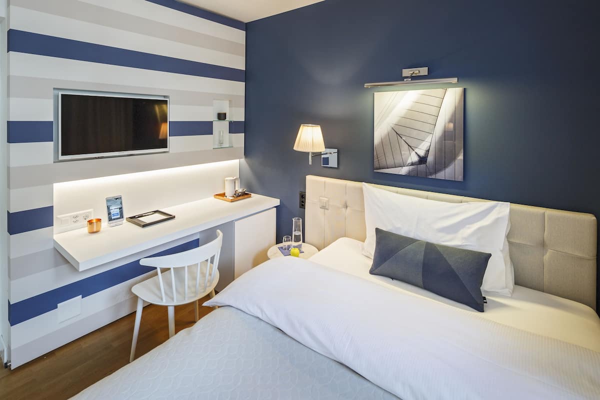 A hotel room with a bed, a desk, a chair, a TV, a lamp, and a painting on the wall.