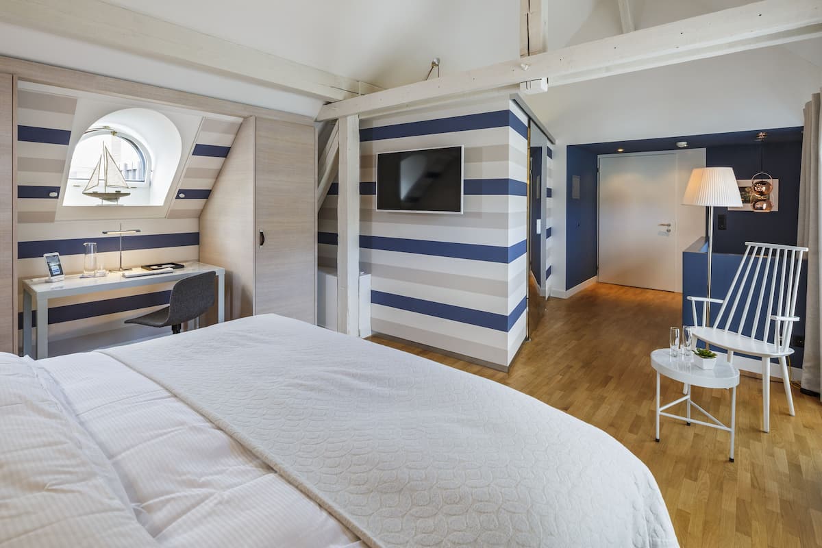 A bedroom with a bed, a desk, a chair, a TV, and a window with a sailboat decoration.