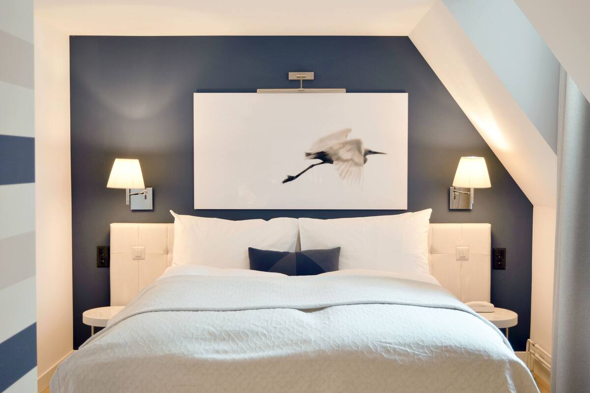 A modern hotel room with a large bed, two bedside tables, a wall-mounted lamp, and a framed picture of a bird.