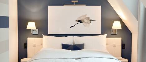 Une chambre d’hôtel moderne avec un grand lit, deux tables de chevet, une lampe fixée au mur et un tableau représentant un oiseau.