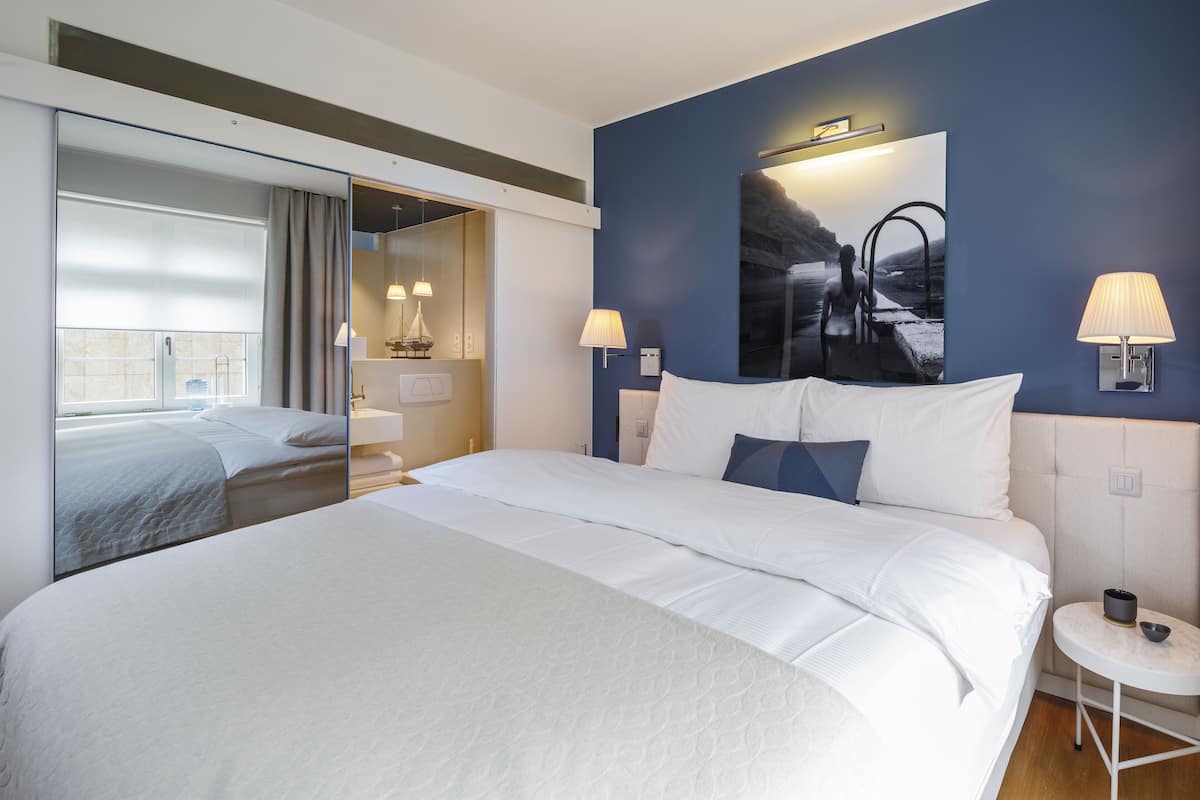 A modern hotel room with a large bed, a mirror, a nightstand, a small table, and a wall-mounted lamp.