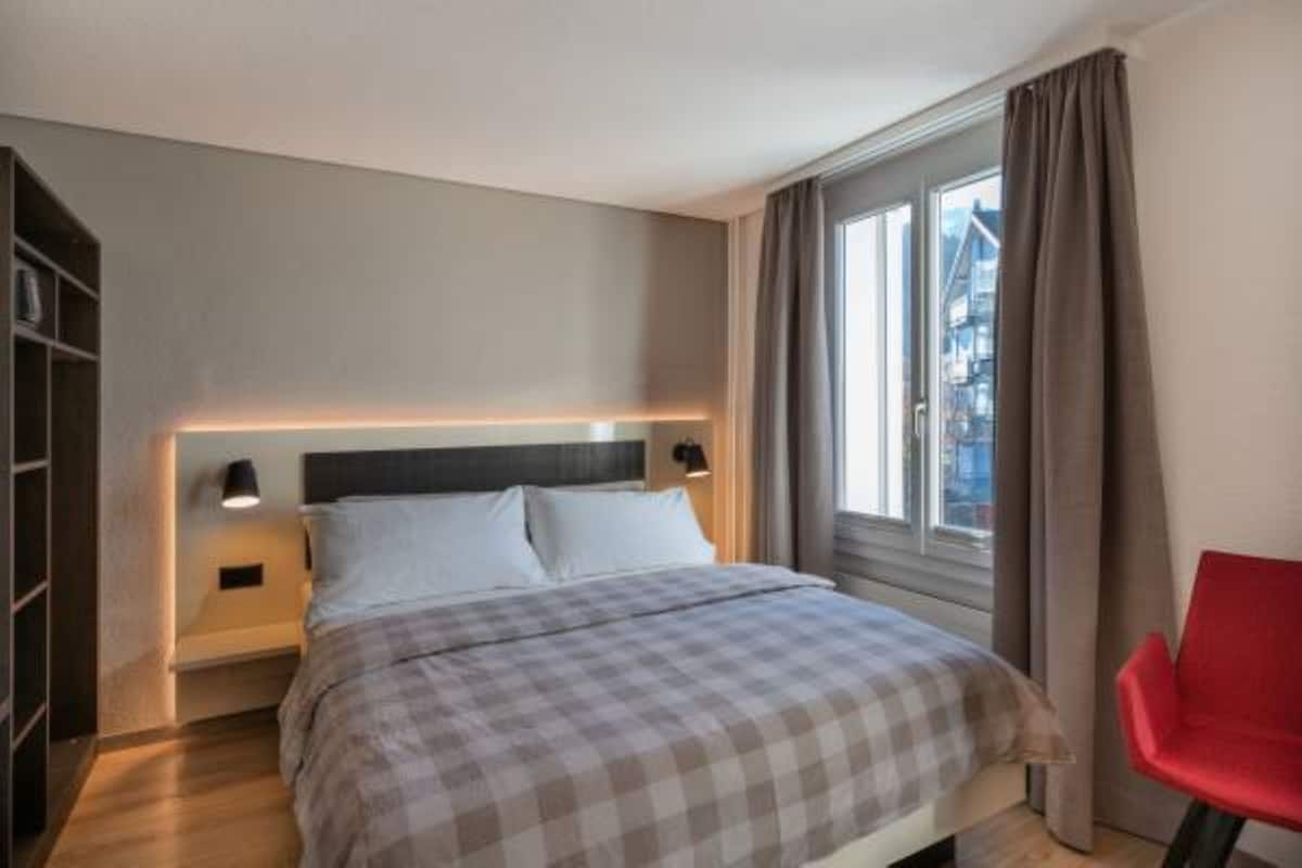 Une chambre à coucher avec un lit, des lampes de chevet, une fenêtre avec des rideaux et un fauteuil rouge.