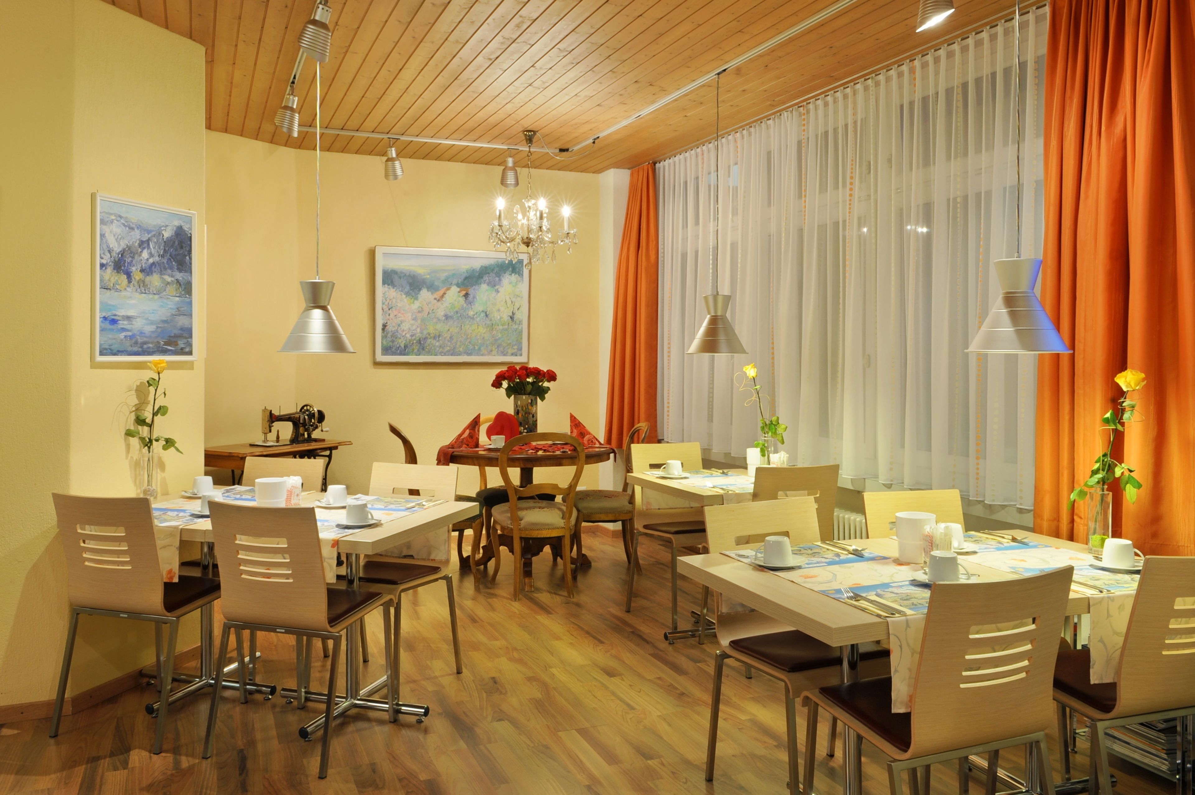 Un comedor con mesas y sillas de madera, y un candelabro. Hay cuadros en las paredes y una máquina de coser sobre una mesa.